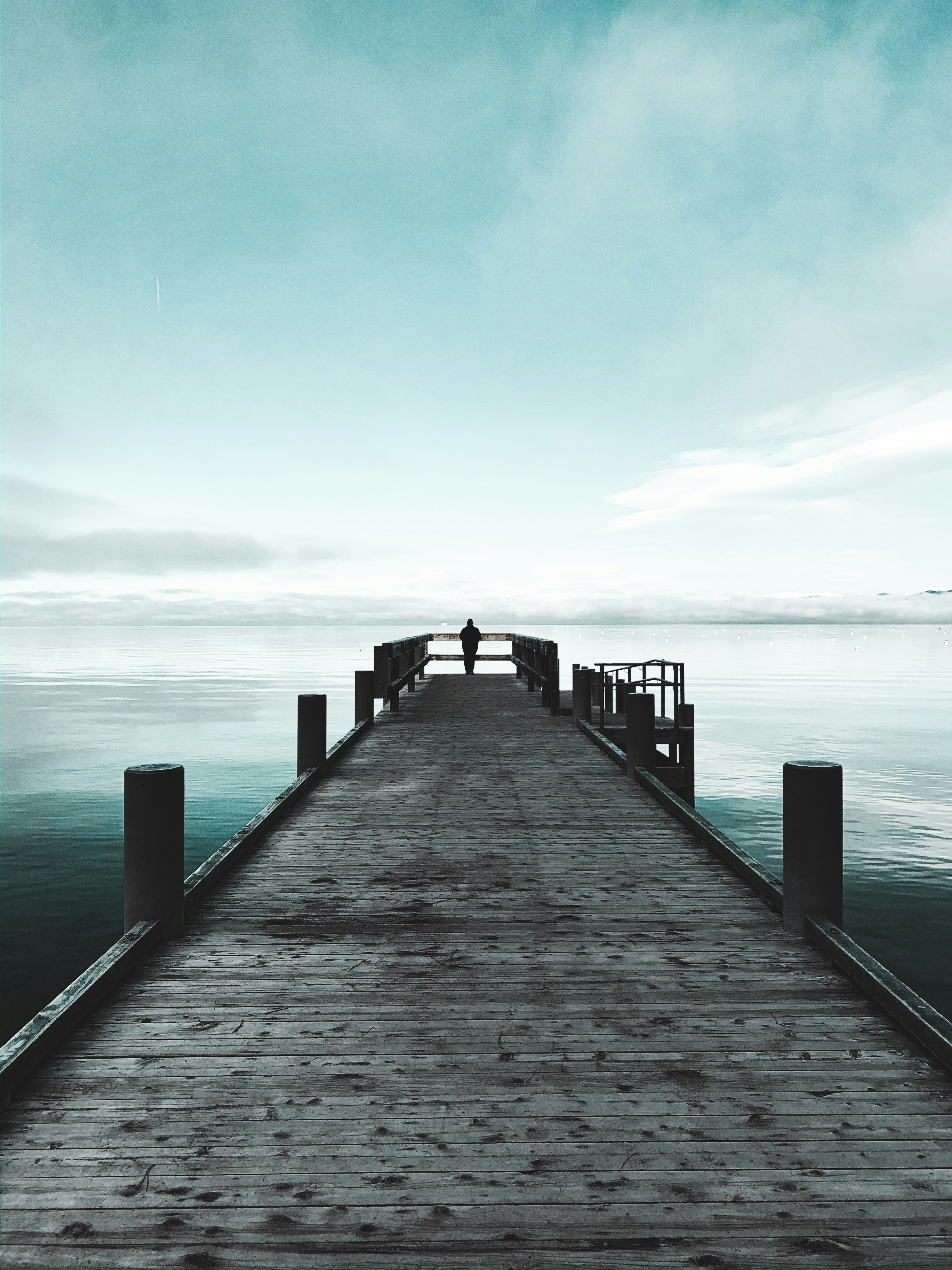 A lone figure standing at the end of a long wooden dock over calm water